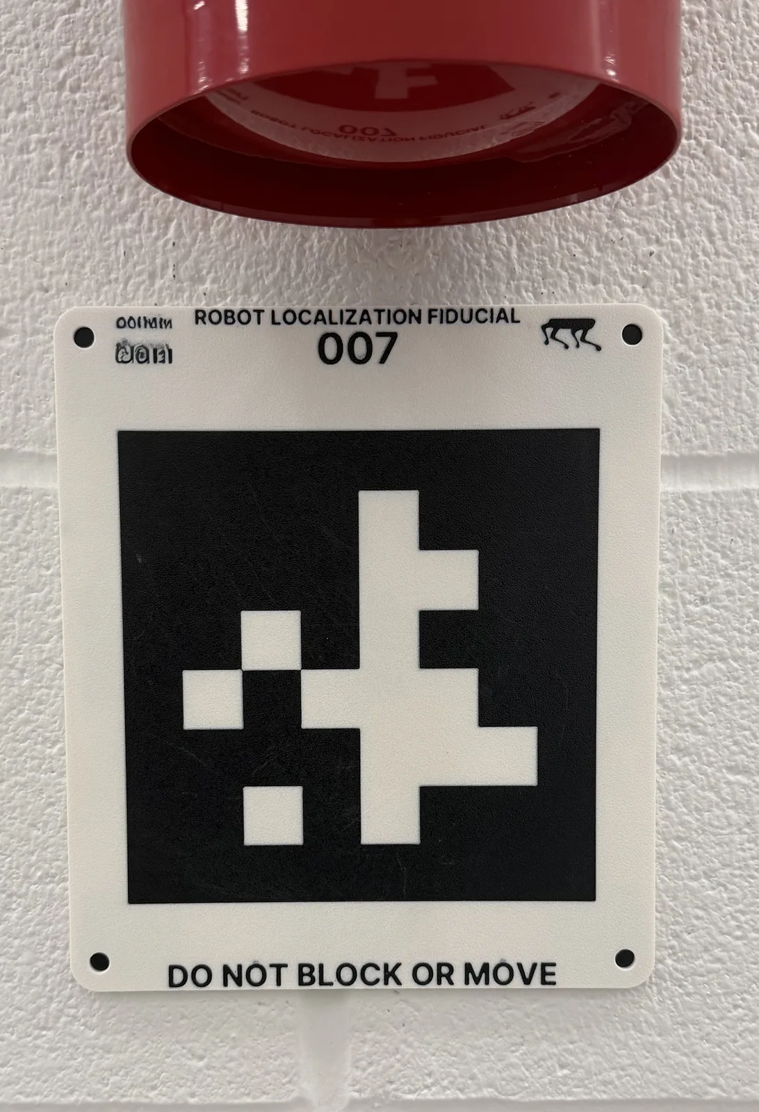 흰색 벽에 고정된 3D 프린트 fiducial plate. 상단에 ROBOT LOCALIZATION FIDUCIAL 007, 하단에 DO NOT BLOCK OR MOVE 문구가 있고 가운데에 검은색 AprilTag가 있다
